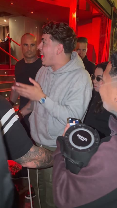 A guy in a grey hoodie steps up to the speaker and raps back at the camera, both hands going, the red stairs behind him.