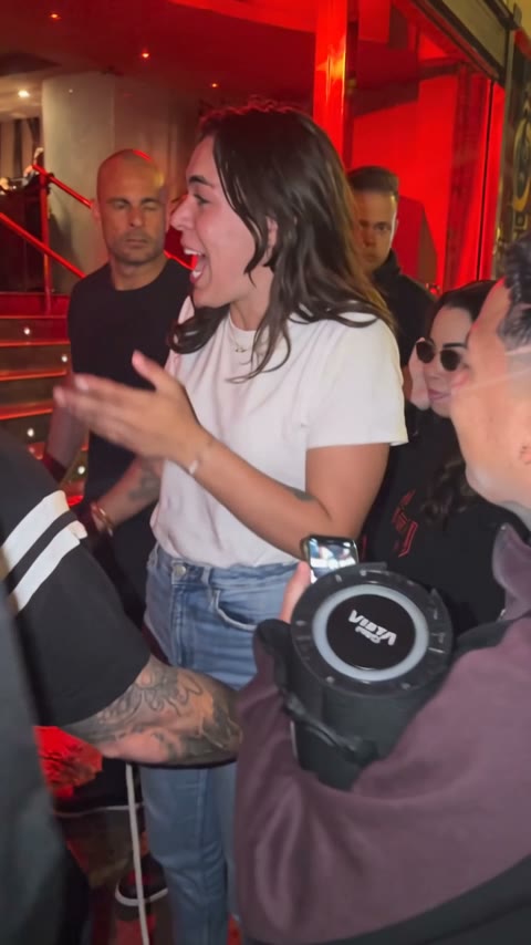 A woman in a white tee takes the freestyle at the red-lit door, laughing and dancing to the beat.