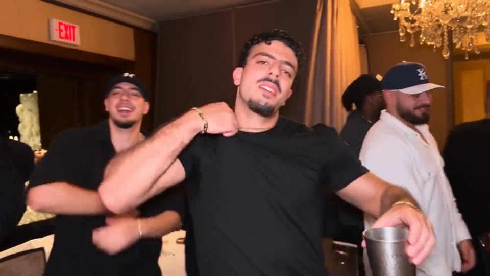 The same moves under the chandeliers of a hotel ballroom, black tee and gold chain.