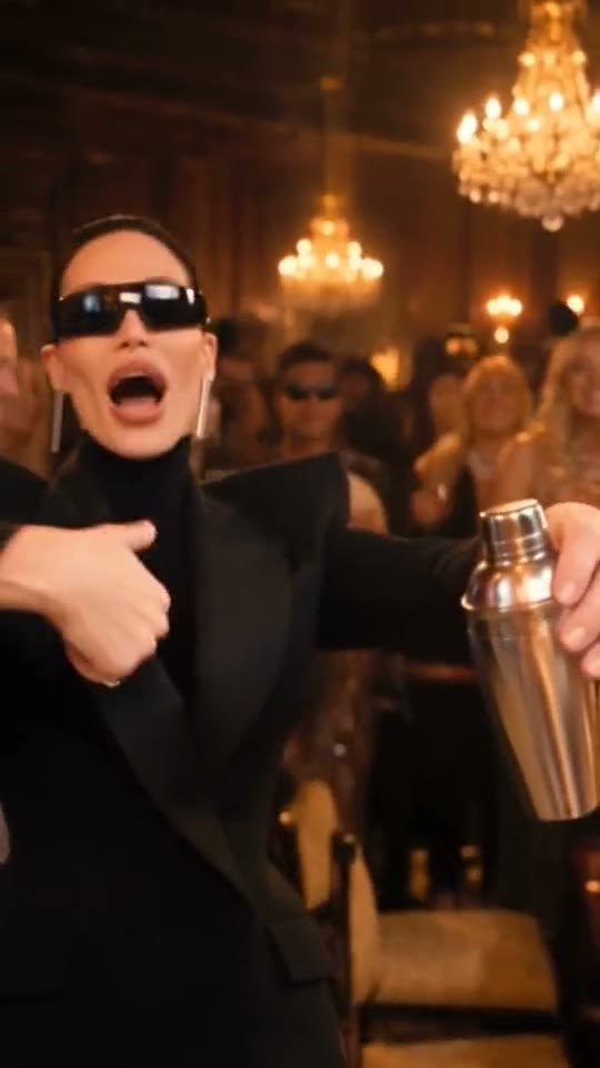 A woman in a black suit and sunglasses takes the lead, shaker in hand.