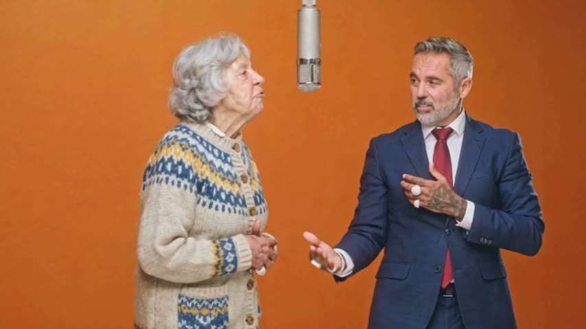 A grandmother in a knitted cardigan on the left and a man in a navy suit on the right run the whole routine in the orange booth.